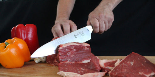 Cuchillo RANTY demostrando técnica profesional de corte en carnes sobre tabla de madera junto a vegetales frescos para preparación.