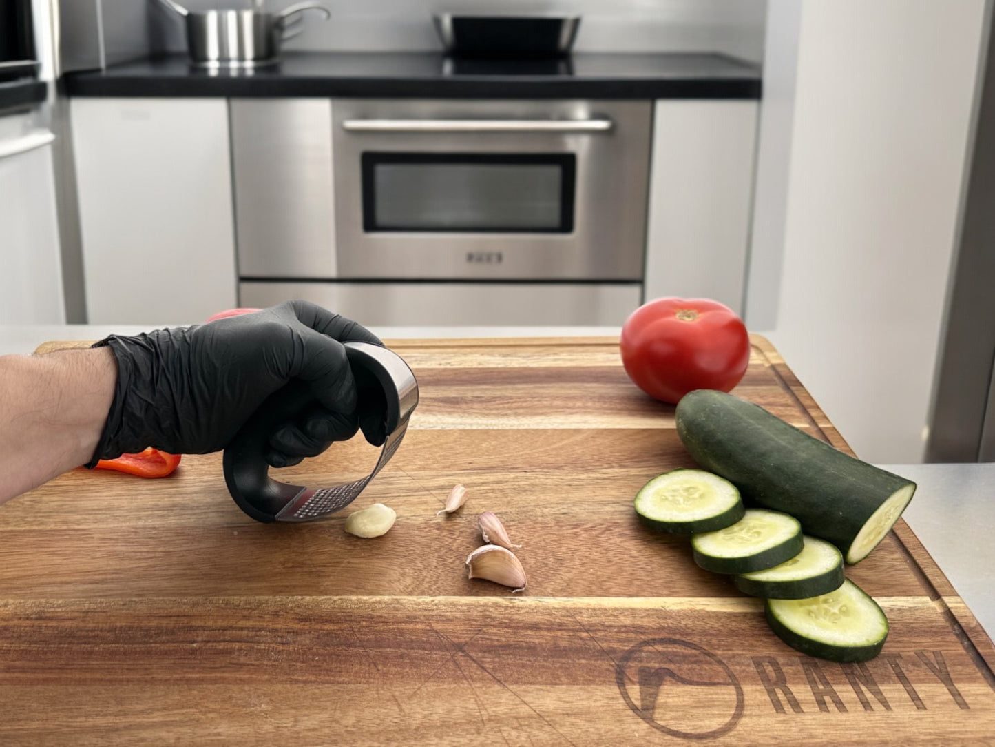 Moledor de ajo balancín Ranty con guante de cocina negro, triturando ajo junto a verduras frescas sobre tabla de madera.