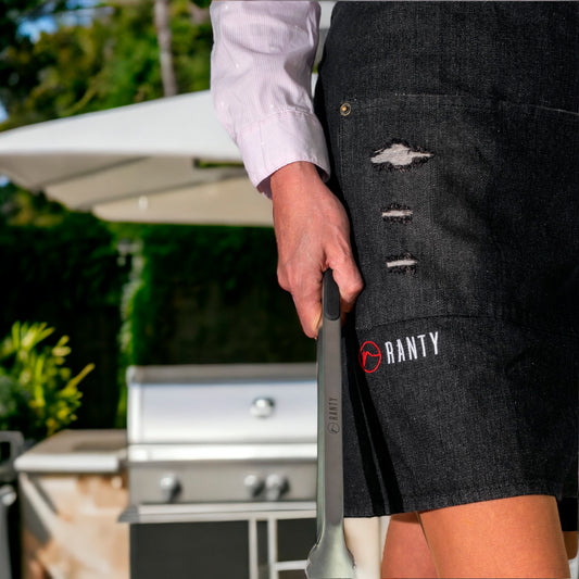 Delantal de cocina vintage jeans Ranty negro con detalles desgastados junto a parrilla exterior.